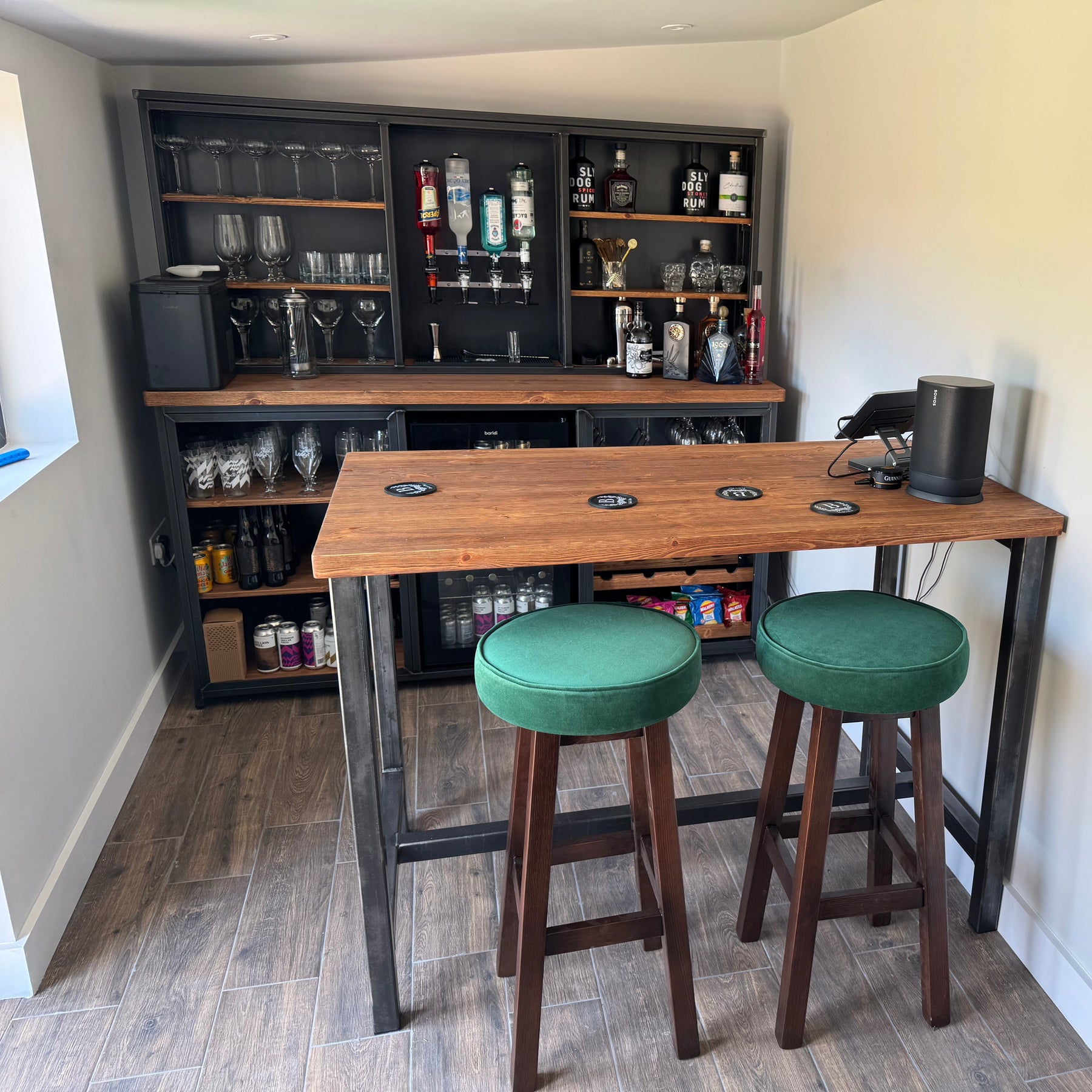 A rustic industrial-style pub bar table with a wooden top and dark metal frame, paired with two green upholstered bar stools. The setup is placed in a room with wood-look flooring, featuring a dark bar cabinet filled with glasses and bottles in the background.
