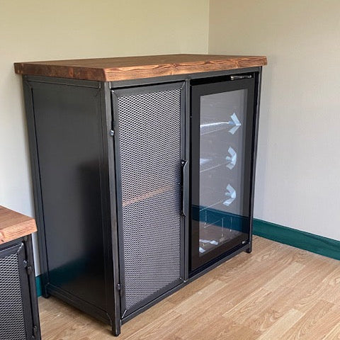 coffee bar cabinet with a rustic wood top, featuring one solid door and one glass door with a black frame, set against a beige wall and light wood floor.