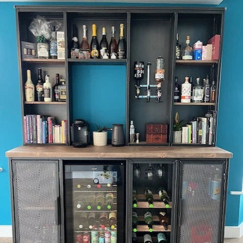 A large black metal bar and wine storage cabinet with an open top shelf section, open side shelves, a wood-look countertop, and two glass-door wine fridges below. The cabinet is filled with bottles, glasses, books, and bar accessories, set against a bright blue wall and light wood floor.