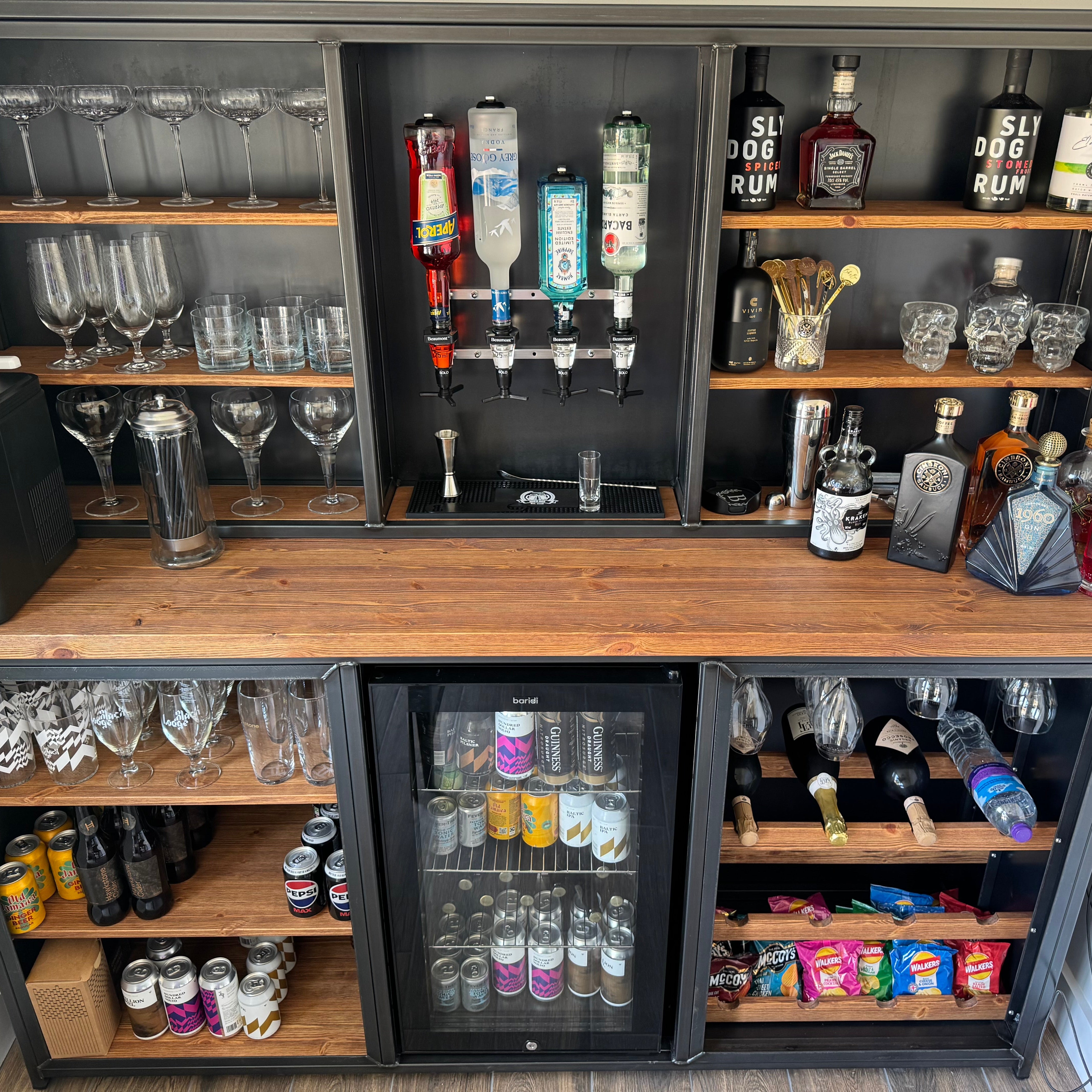 A large black and wood bar cart or home bar cabinet filled with bottles, glasses, and bar accessories, photographed indoors against a plain wall.