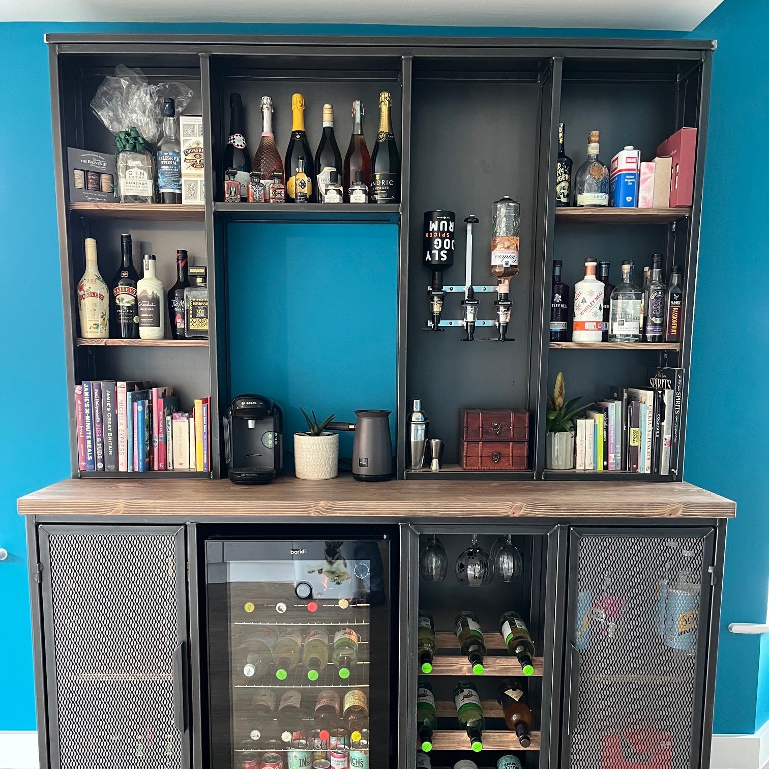 A large industrial-style bar and beverage storage unit with black metal shelving, wood-look side panels, mesh doors, and a glass-door refrigerator section filled with bottled drinks. The unit is shown against a blue wall and light wood floor.