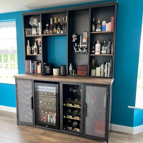 A tall black metal and wood bar cart with multiple shelves, mesh-front storage compartments, and a glass-door wine cooler, shown in a room with blue walls and a wood floor.